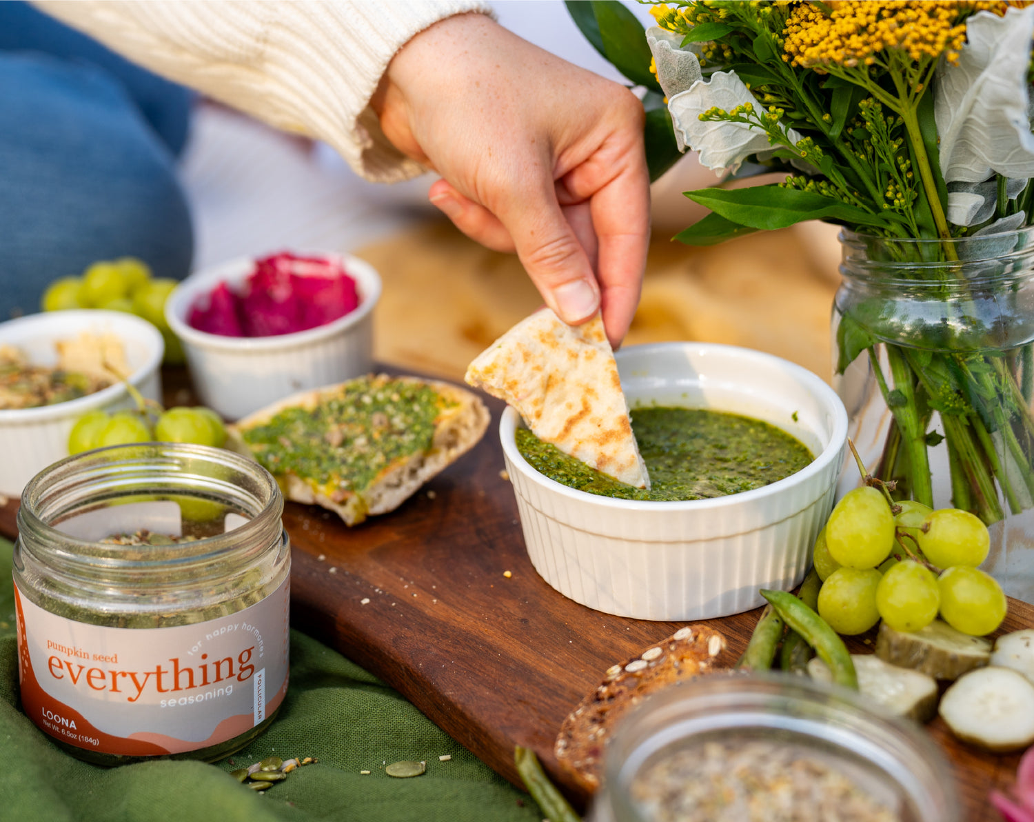 A charcuterie board of pita and other bread with pesto and beet spreads, sprinkled with Loona Seed blends. A hand dipping pita into pesto.