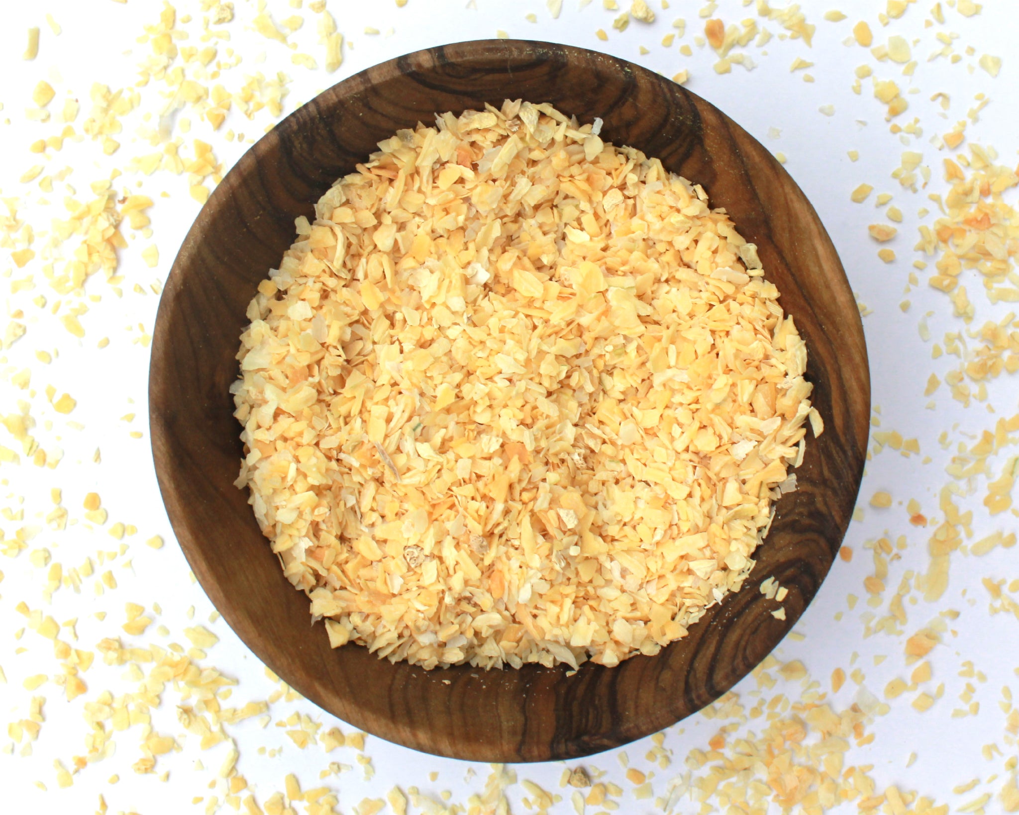 A small wooden bowl filled with garlic and onion granules, on a white background with granules scattered around it.