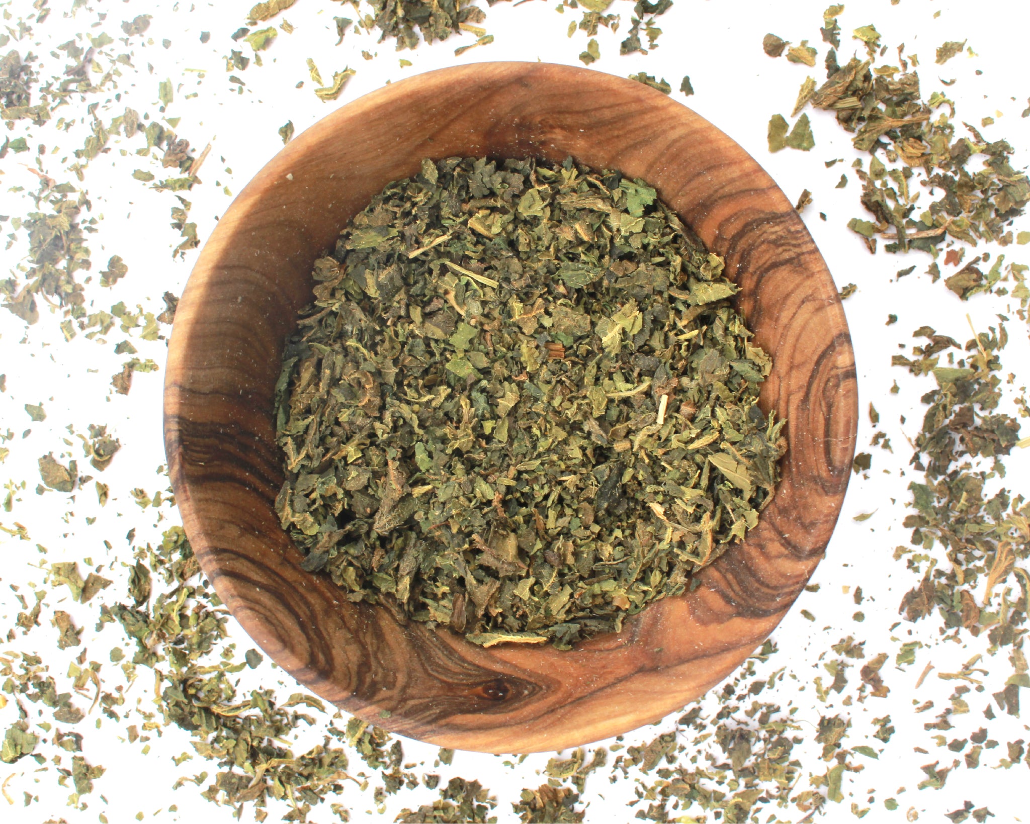 A small wooden bowl filled with nettle leaf, on a white background with leaves scattered around it.