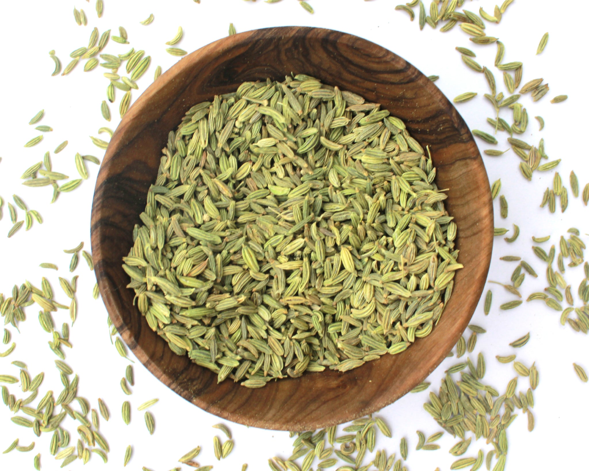 A small wooden bowl filled with fennel seeds, on a white background with seeds scattered around it.