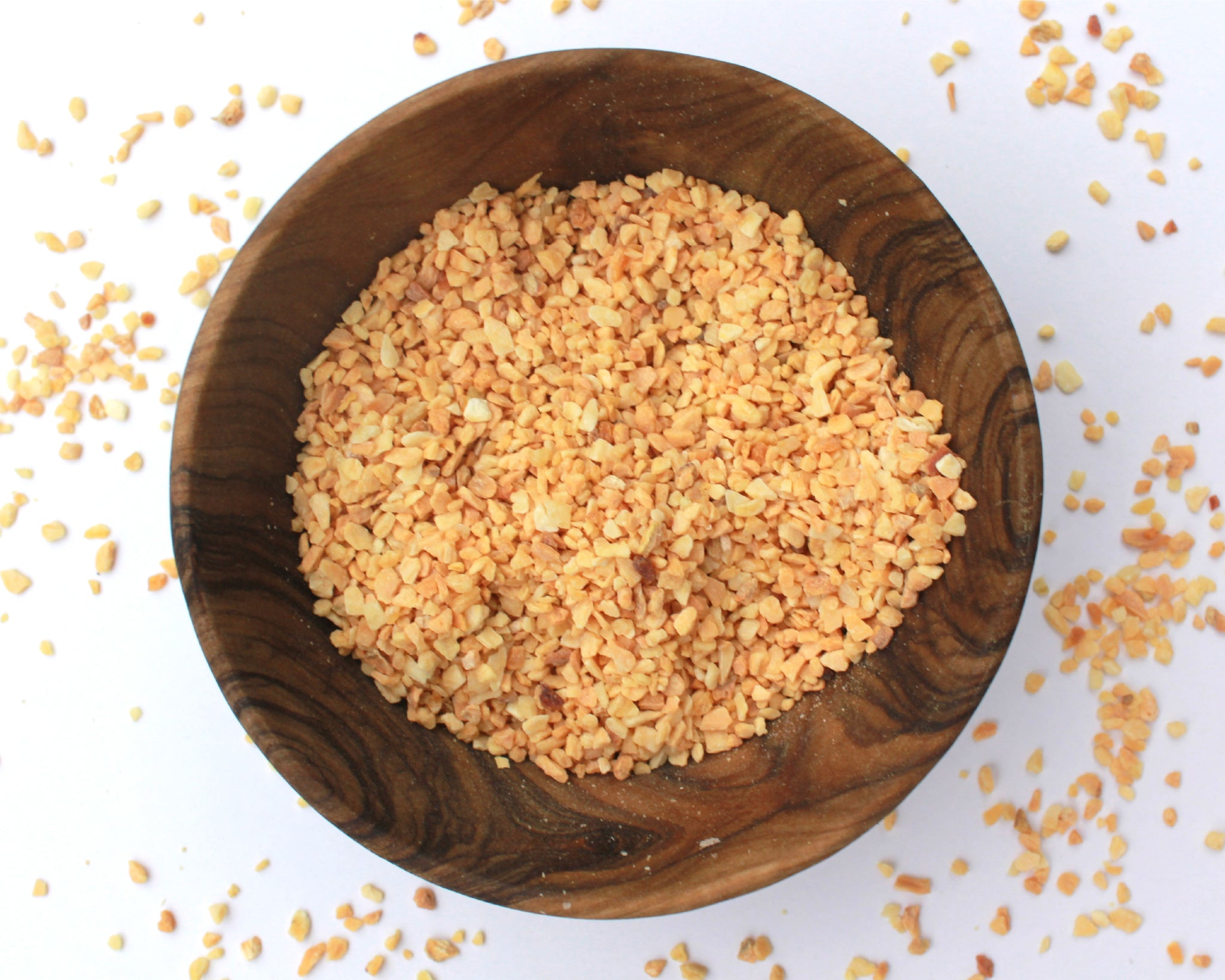 A small wooden bowl filled with fenugreek seeds, on a white background with seeds scattered around it.