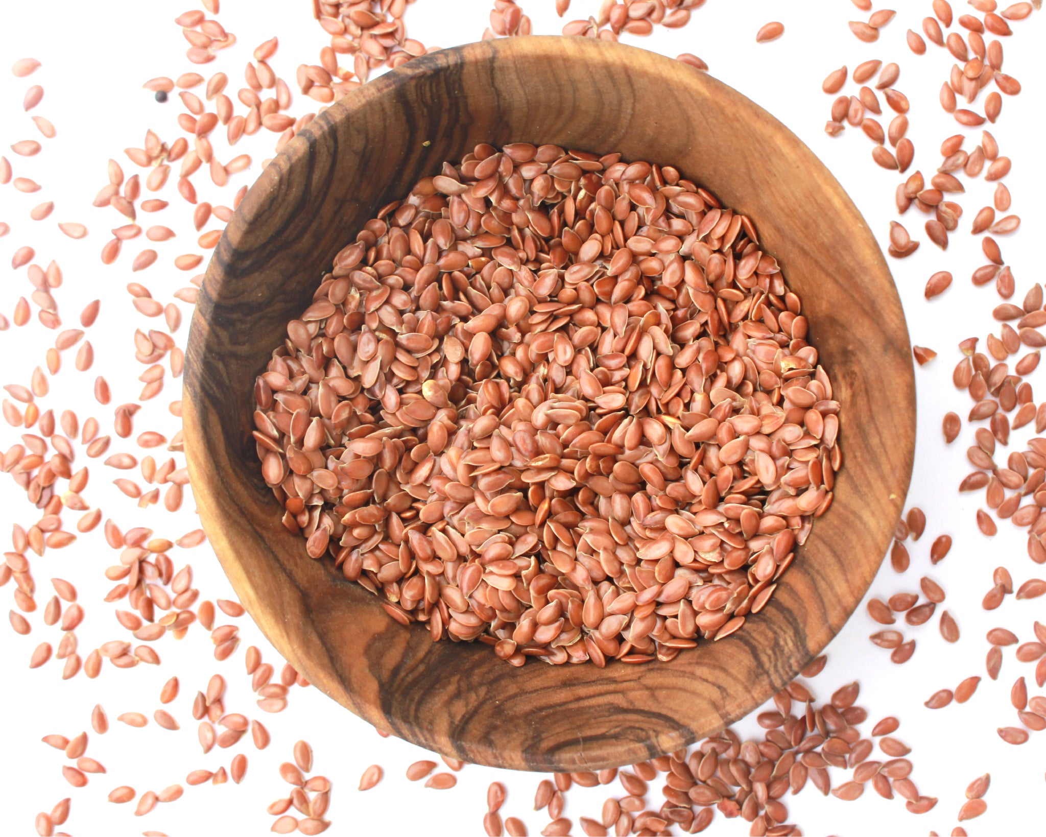 A small wooden bowl filled with flax seeds, on a white background with seeds scattered around it.