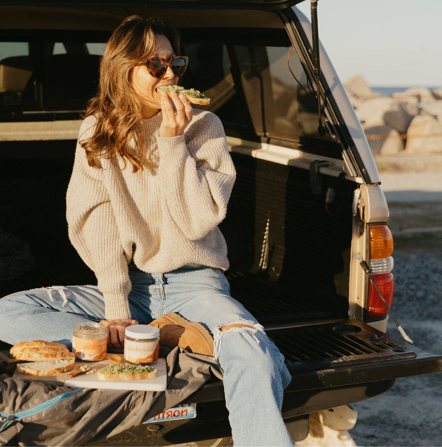 A woman in sunglasses, dressed in a sweater and jeans, sitting in an open hatchback back seat, eating a piece of toast with seeds sprinkled on top.