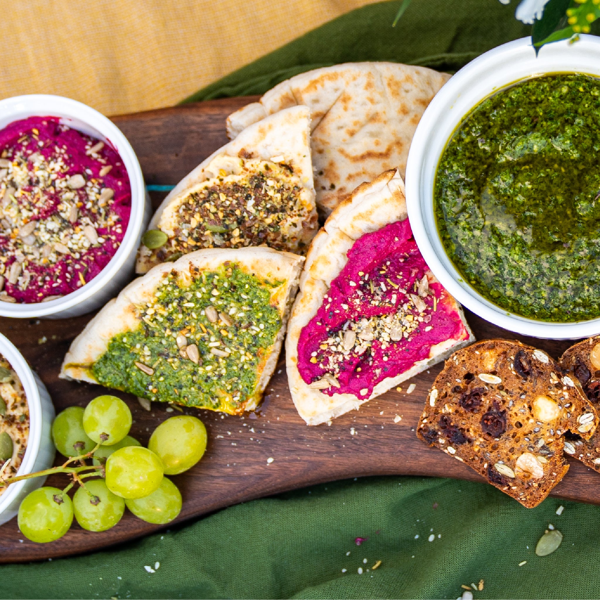 A charcuterie board of pita and other bread with pesto and beet spreads, sprinkled with Loona Seed blends.