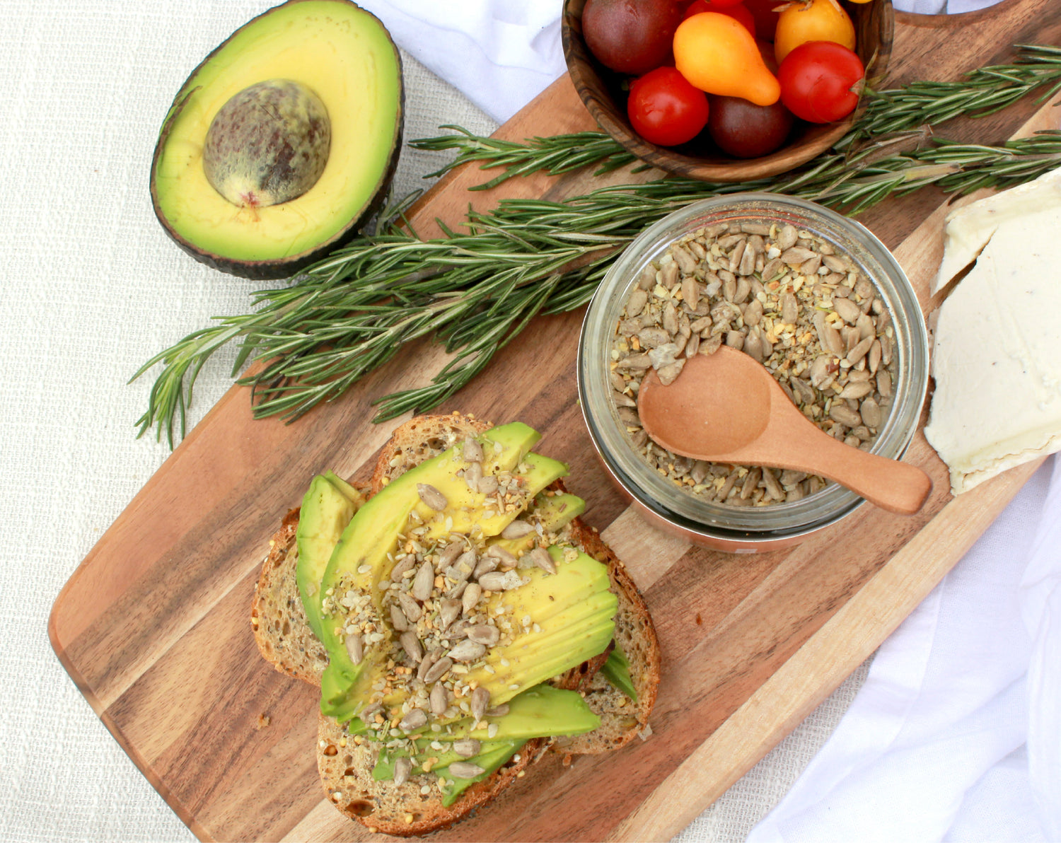 A charcuterie board of avocado, grape tomatoes, rosemary, and toast sprinkled with seeds.