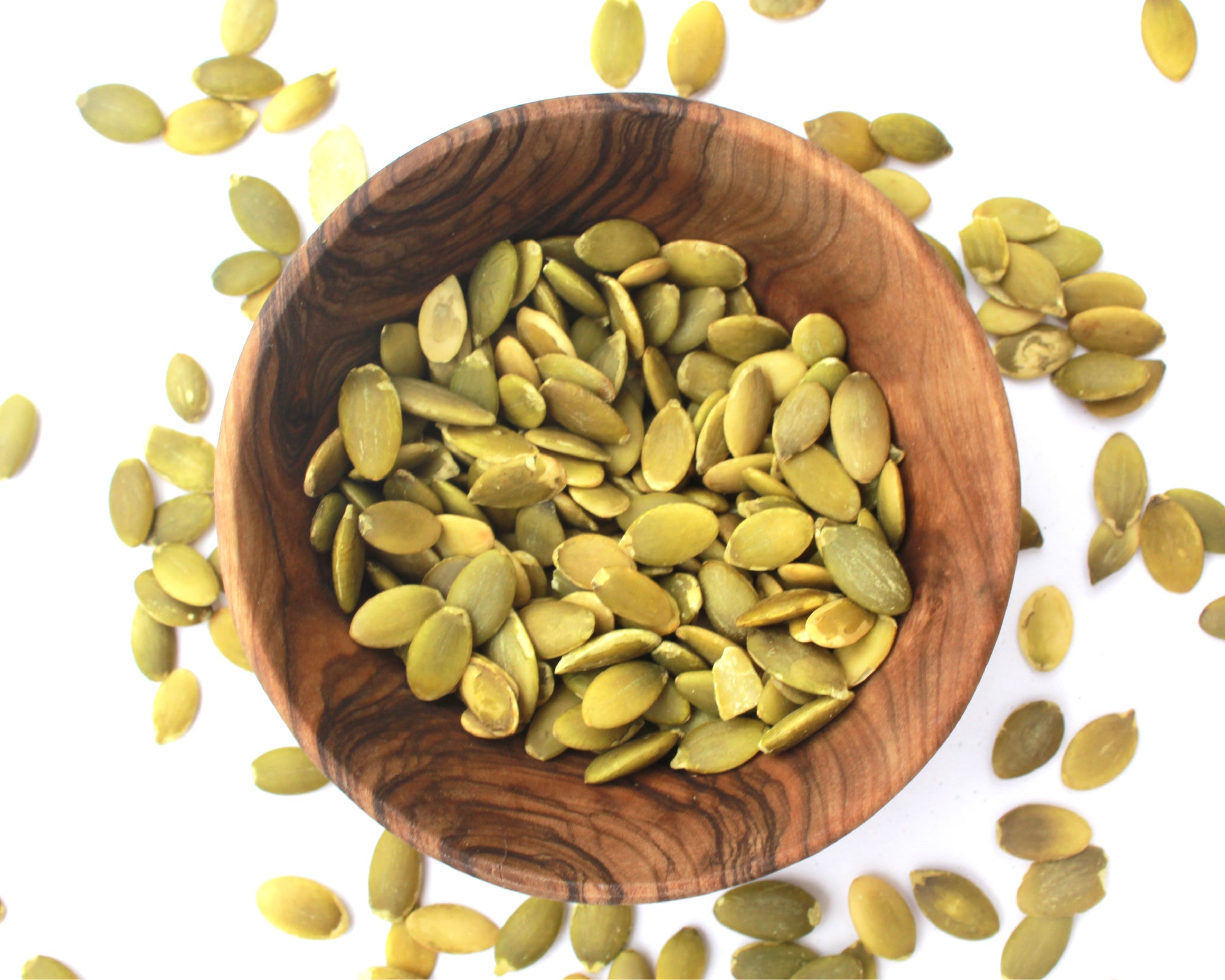 A small wooden bowl filled with pumpkin seeds, on a white background with seeds scattered around it.