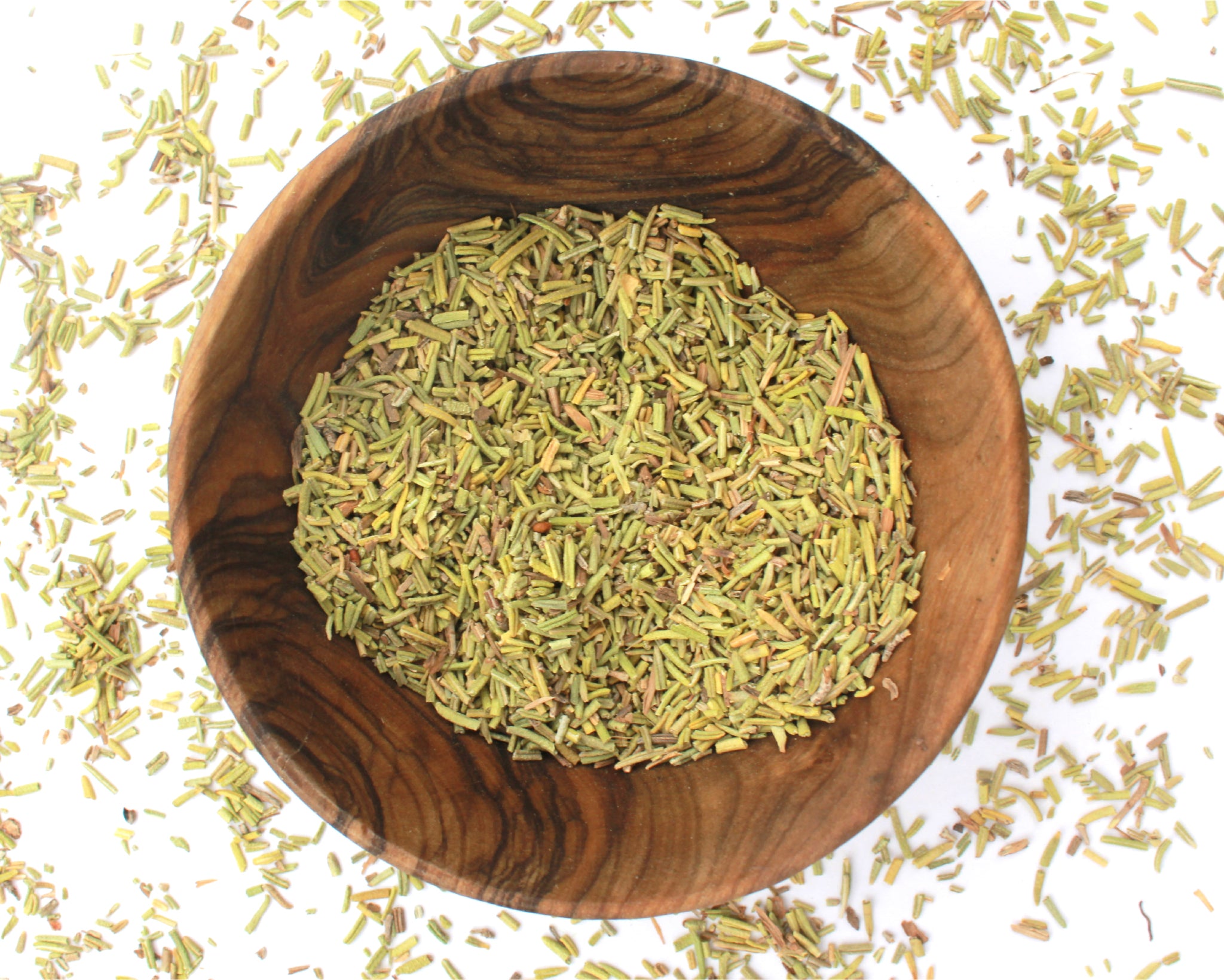 A small wooden bowl filled with rosemary leaf, on a white background with leaves scattered around it.