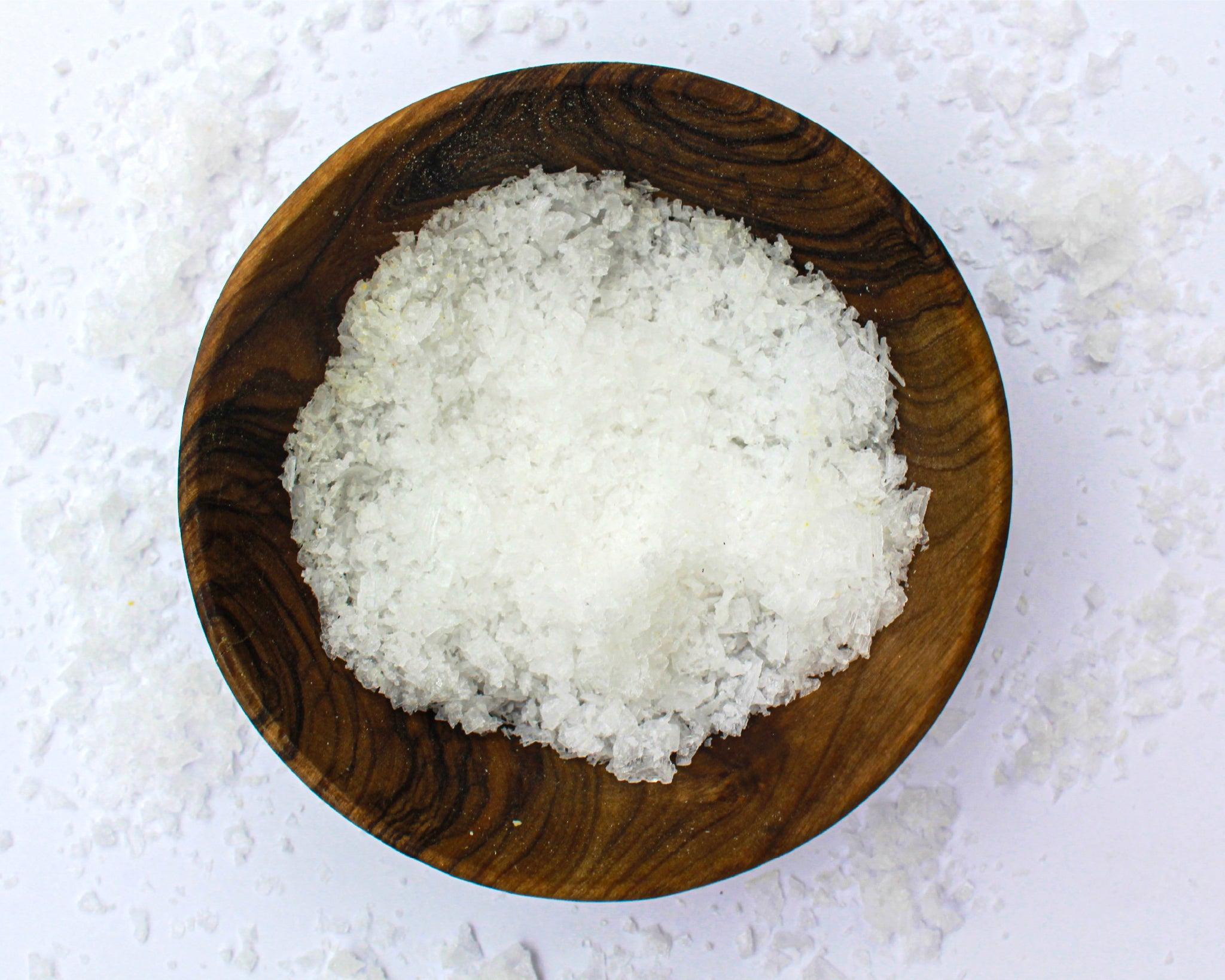 A small wooden bowl filled with sea salt, on a white background with salt scattered around it.