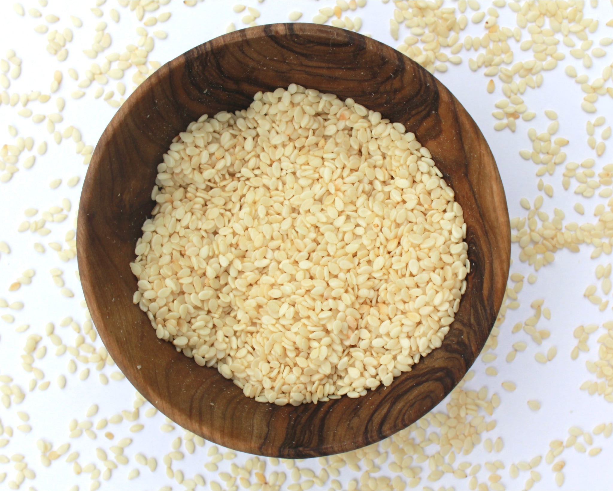 A small wooden bowl filled with sesame seeds, on a white background with seeds scattered around it.