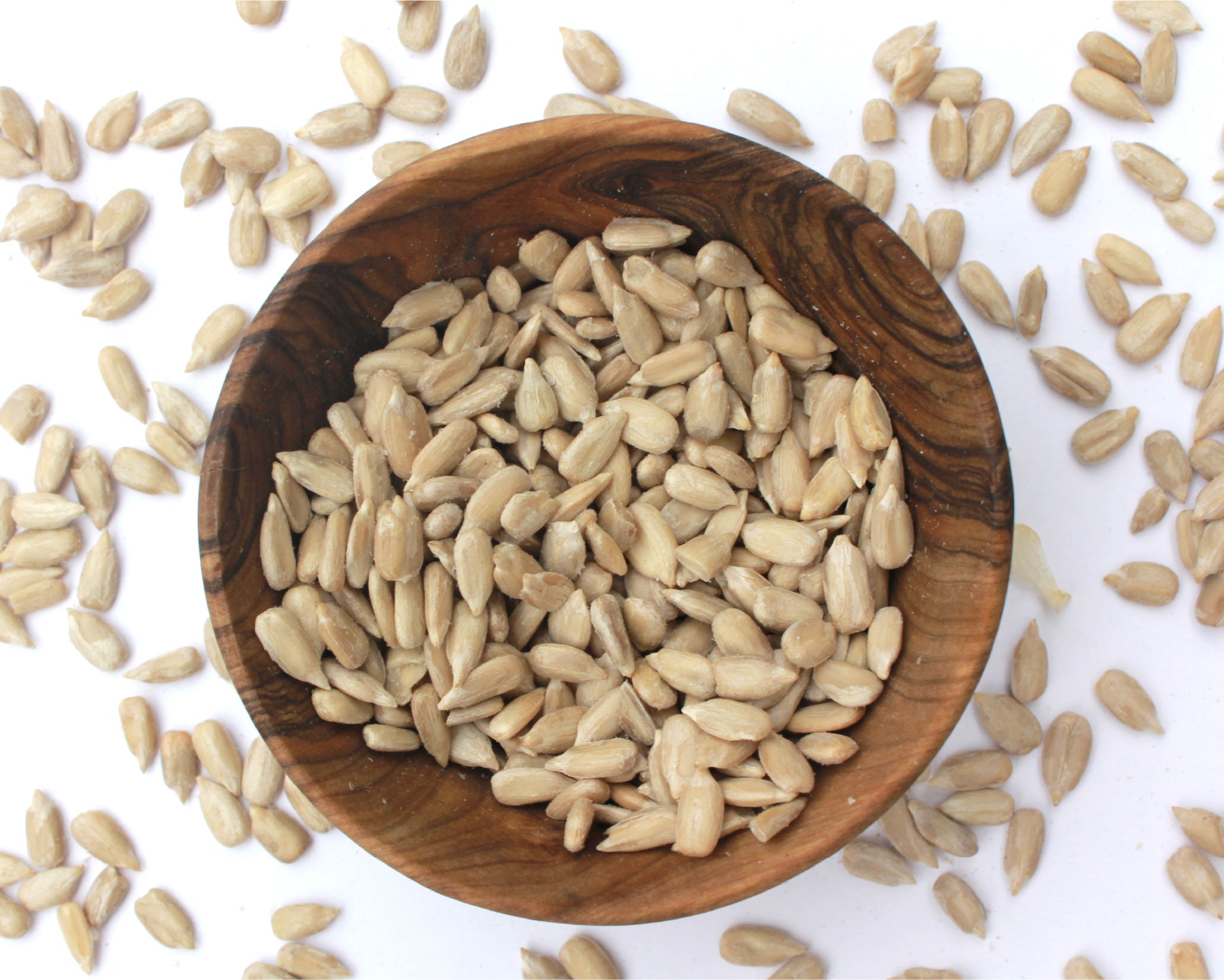 A small wooden bowl filled with sunflower seeds, on a white background with seeds scattered around it.